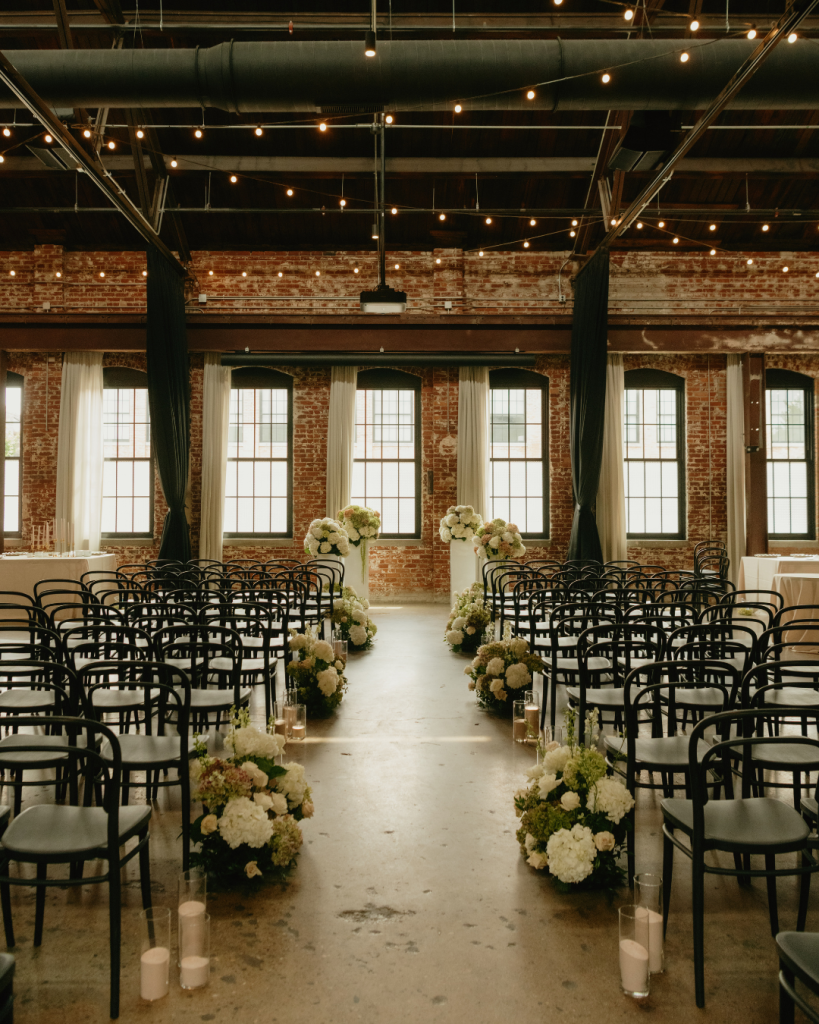 Indoor wedding ceremony at The Ellis with black chairs, exposed brick walls, tall windows, candles, and aisle florals.