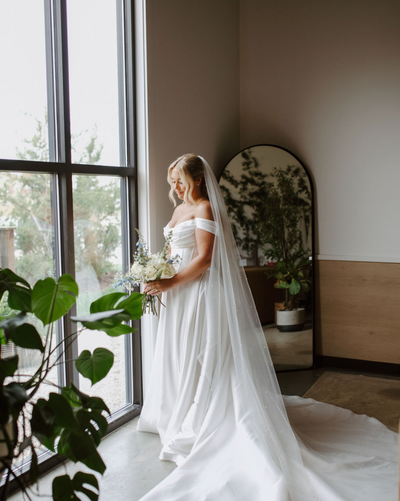 Bride Lainey stands beside a large window at Retreat 21 holding a white and dusty blue bouquet in her off-the-shoulder wedding gown.
