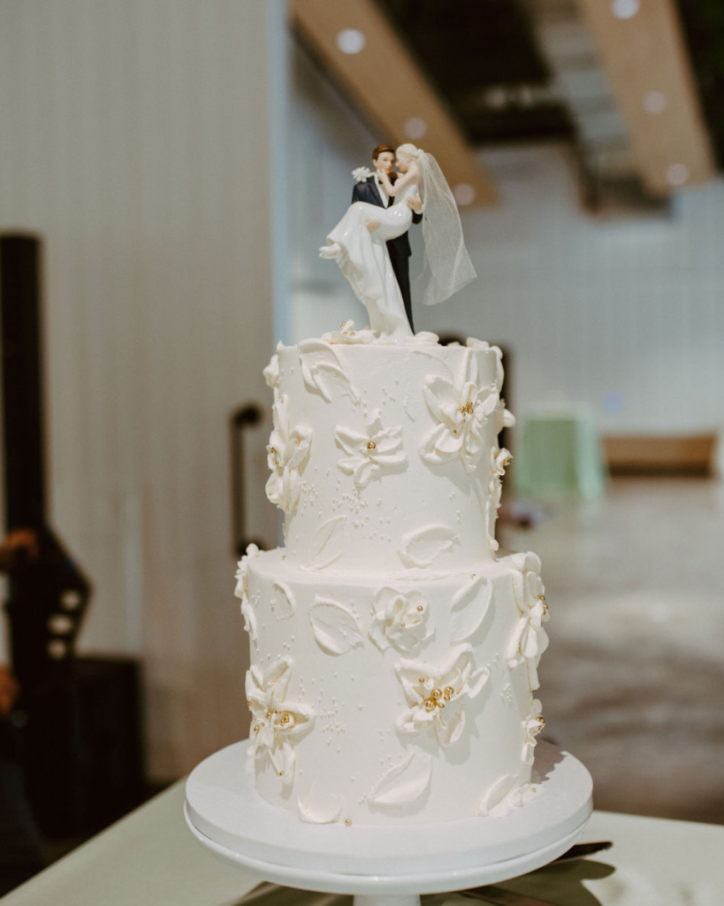 Two-tier white wedding cake decorated with sculpted flowers, gold accents and a classic bride-and-groom topper.