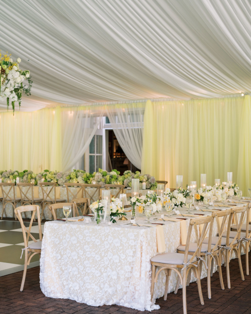 Tented reception features pale yellow draping, floral linens, cross-back chairs, candles and white flowers.