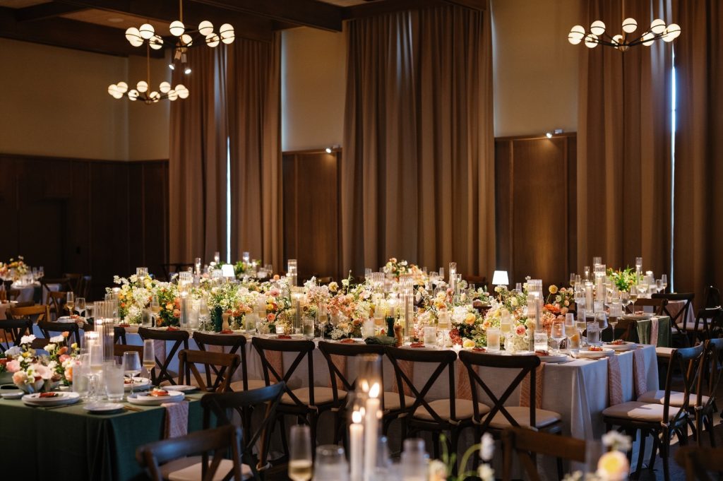Elegant indoor wedding reception with candlelit tables, tall floral centerpieces and modern chandeliers creating a warm, intimate atmosphere.