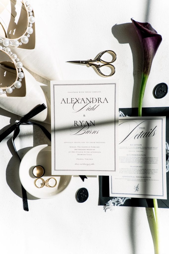 Mackenzie Grace Design wedding invitation and details card styled with pearl-trimmed heels, rings, a dark calla lily, and wax seals.