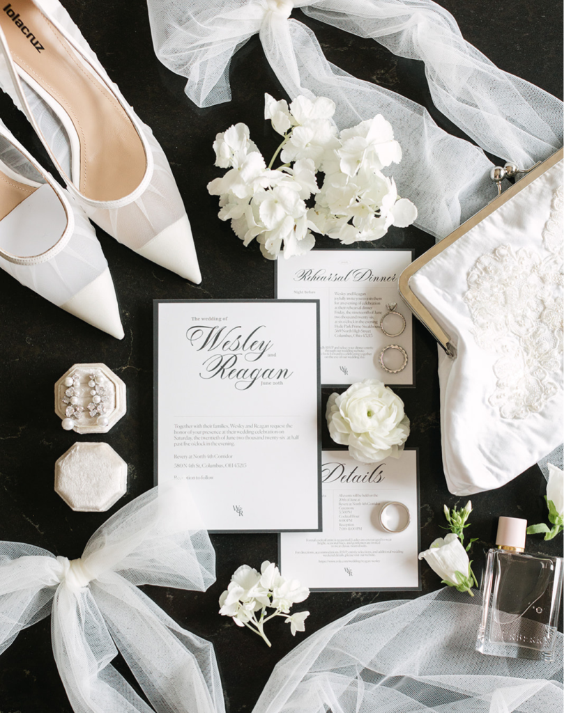 Reagan and Wes’s wedding stationery displayed with white shoes, jewelry, flowers, perfume and bridal accessories.