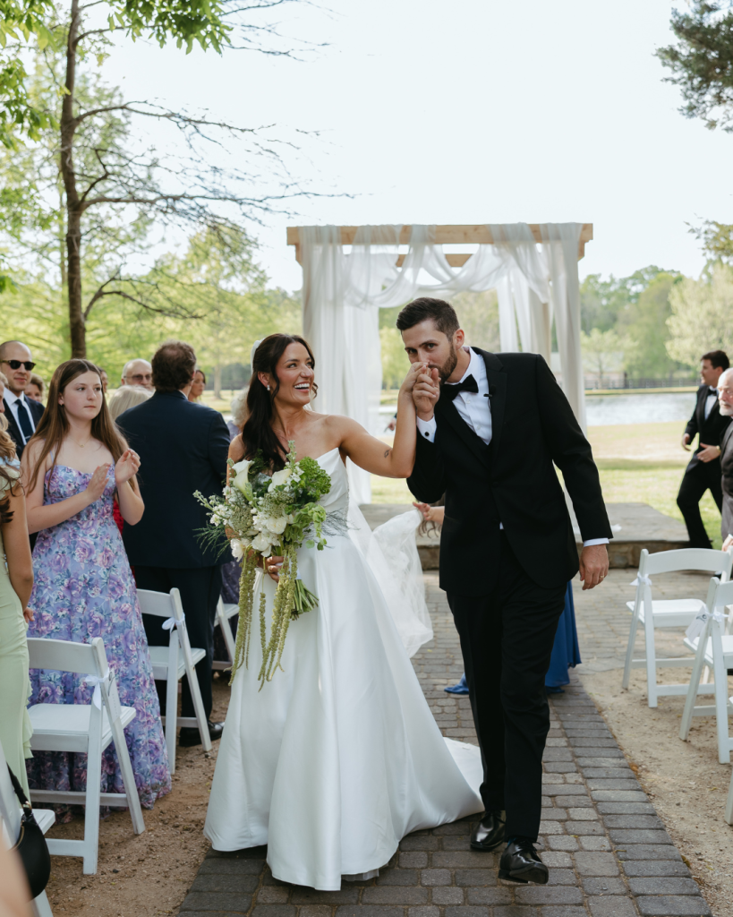 Newlyweds Selina and Sam walk down the aisle after their outdoor spring wedding ceremony at Morning Glory Farm in Monroe, North Carolina, as Sam kisses Selina’s hand.