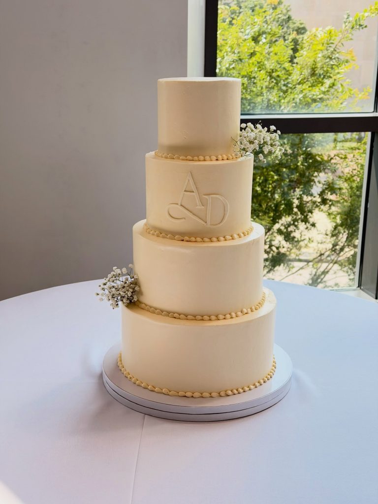 Four-tier white wedding cake with a monogram, delicate gold trim, and baby’s breath.