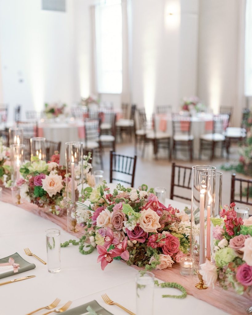 Pink and green layered flowers and pink candles with a pink table runner on a white table for a wedding