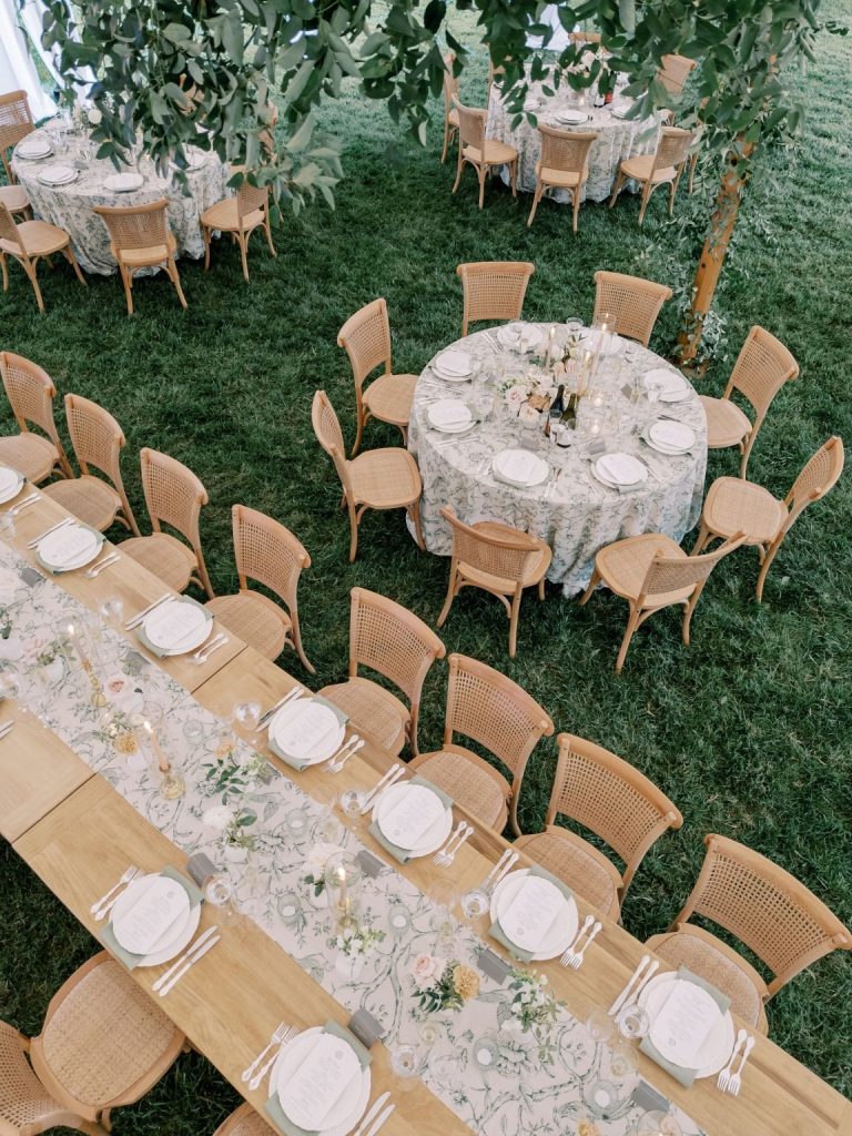 Overhead view of an outdoor wedding reception with long wooden and round tables, floral-patterned linens, soft floral centerpieces, candlelight and natural cane-back chairs arranged on a lush lawn beneath greenery.