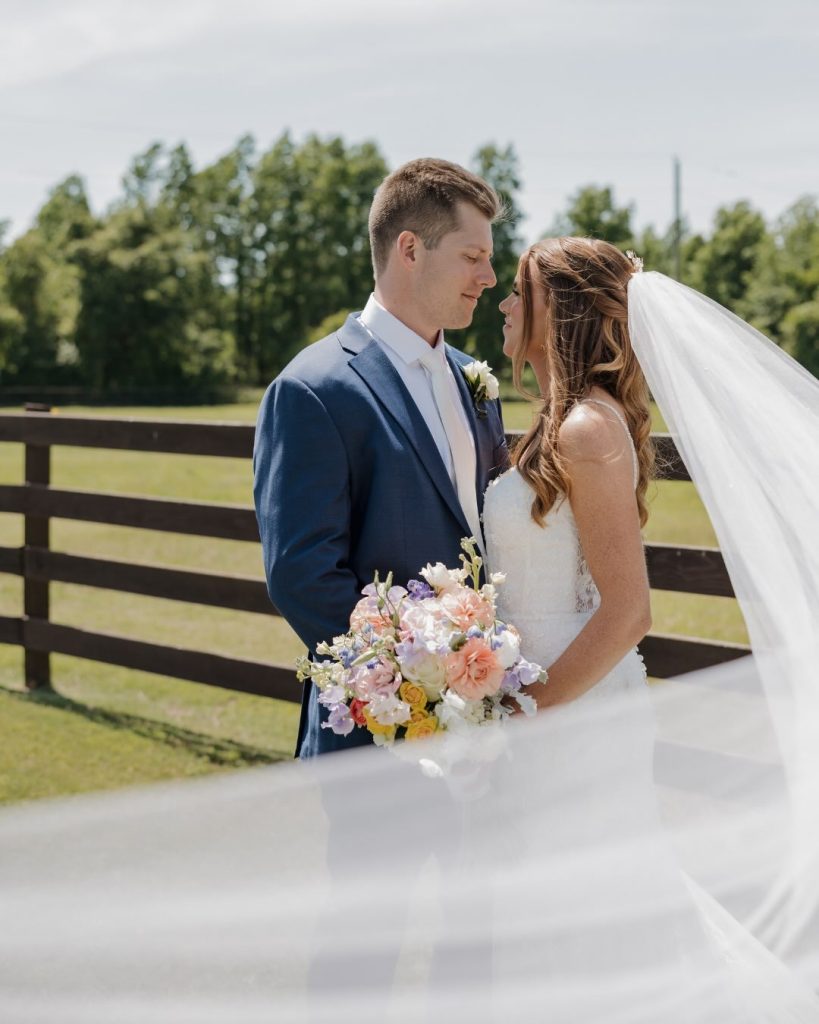 The groom in a navy suit and the bride in a white gown stand close together in front of a dark wooden fence and green pasture. Her veil blows out to the side as she holds a pastel bouquet.