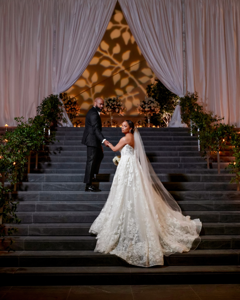 Joy and Edward pose hand in hand on the dramatic staircase at The Ritz Carlton Charlotte, surrounded by greenery, candlelight and white florals with a botanical light projection overhead.