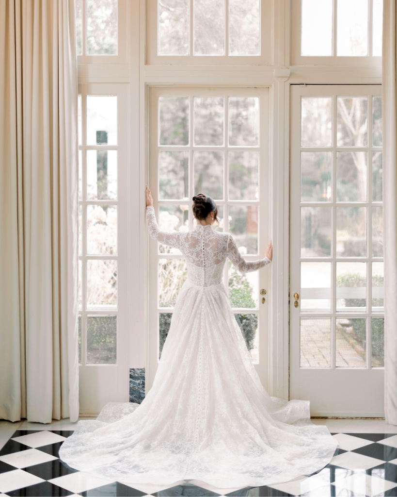 Bride Hannah stands in front of tall windows wearing a long sleeve lace wedding gown with a dramatic train on a black and white checkered floor.