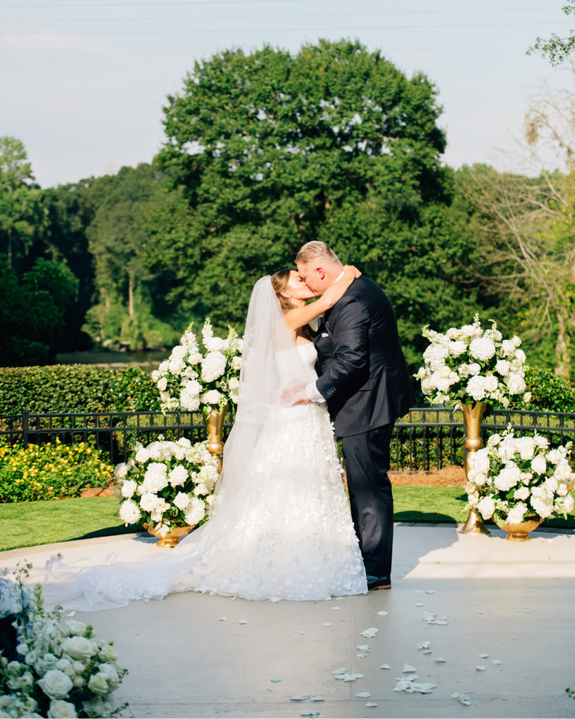 Kelly and Jason kiss at their outdoor wedding ceremony at Riverwalk Event Venue in Rock Hill, South Carolina, surrounded by lush white floral arrangements and greenery.