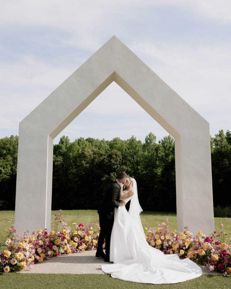 Carly and JT share a kiss beneath the house shaped ceremony arch at The Lumen House, surrounded by colorful spring florals in pink, orange, yellow and blue.