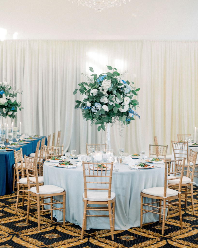 Blue and white wedding reception with tall floral centerpieces, light blue linens, gold chairs, and candlelight.