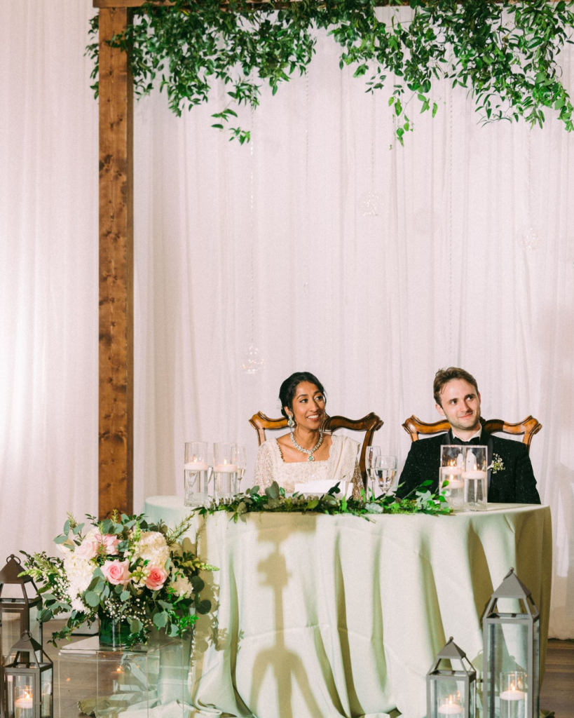 bride and groom sitting at sweetheart table with greenery above them