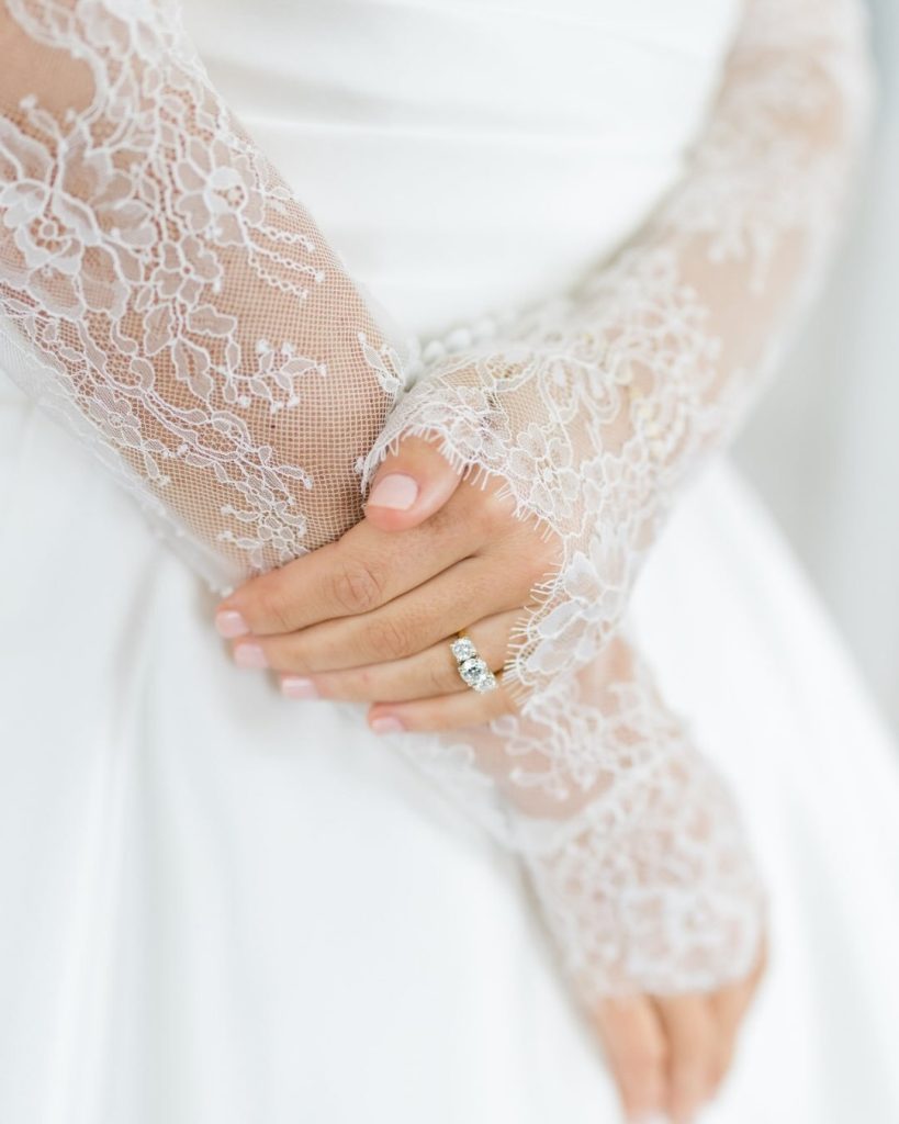 Close-up detail shot of a bride's hands featuring sheer, intricate white lace fingerless gloves and a sparkling diamond engagement ring against her white gown.