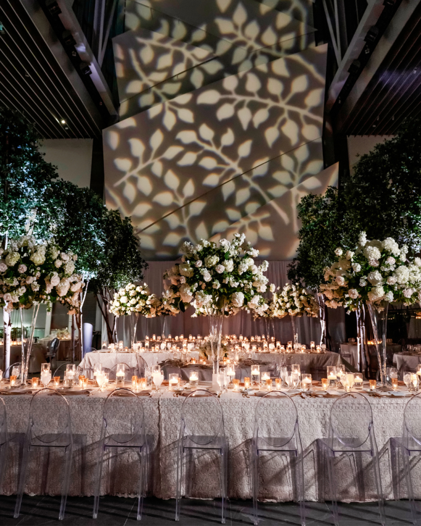 Luxury wedding reception at The Ritz Carlton Charlotte featuring tall white floral arrangements, clear chairs, textured linens, candlelight and large indoor trees.