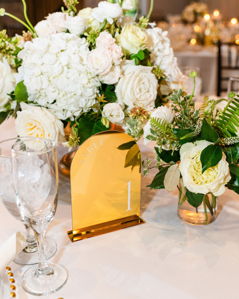 Gold table number one sits beside arrangements of white hydrangeas, roses and greenery at Kelly and Jason’s Riverwalk Event Venue reception.