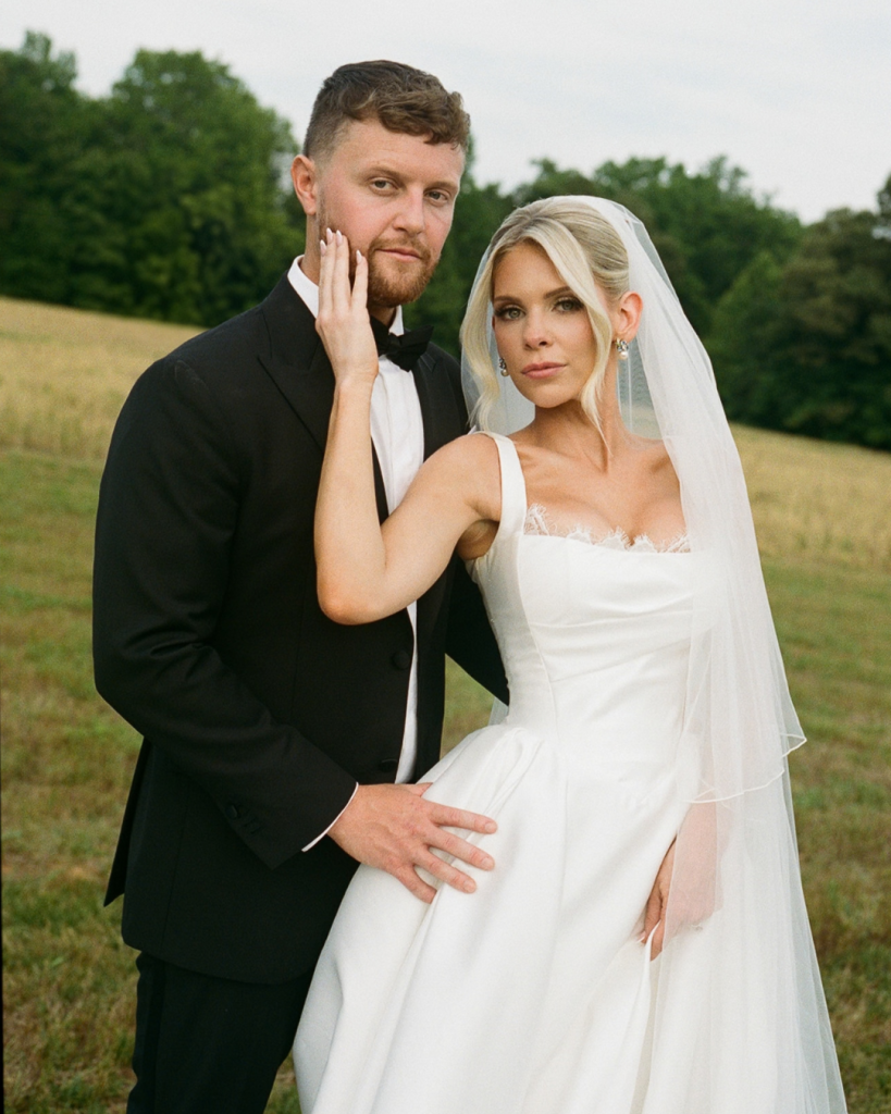 Carly and JT smile together during their wedding ceremony at The Lumen House, with vibrant floral arrangements framing the white house shaped arch behind them.