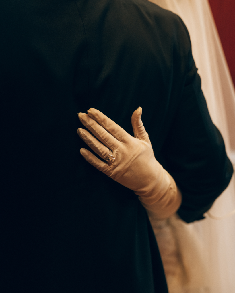 close up of brides rings on sheer gloved hand behind husbands back
