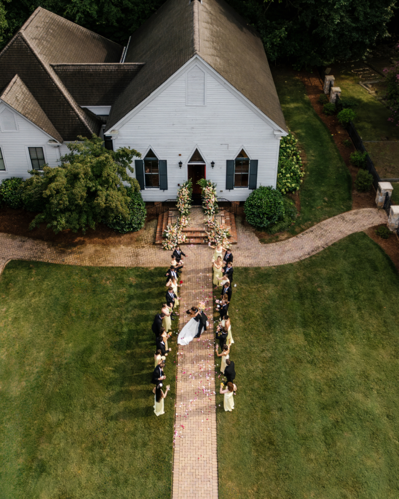 Aerial view of Lauren and Peyton with their wedding party outside the Chapel at Morning Star, surrounded by colorful florals and scattered flower petals.