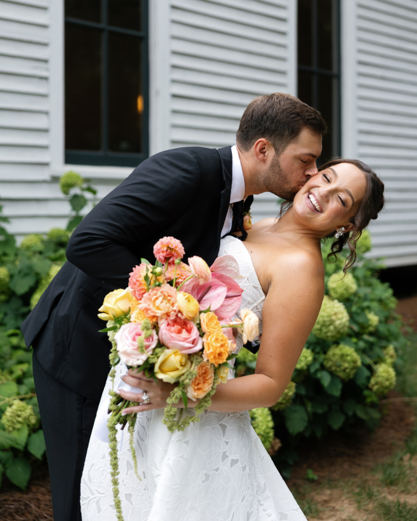 Peyton kisses Lauren on the cheek as she holds a colorful pink, peach and yellow bridal bouquet outside the Chapel at Morning Star.