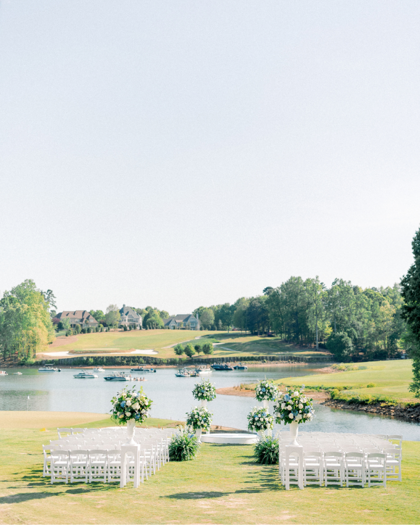 Waterfront wedding ceremony setup with white chairs and large blue, white, and green floral arrangements overlooking the lake.