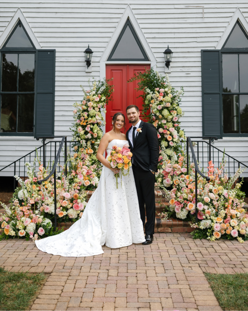 Lauren and Peyton pose outside the Chapel at Morning Star surrounded by abundant pink, peach, yellow and white wedding florals.