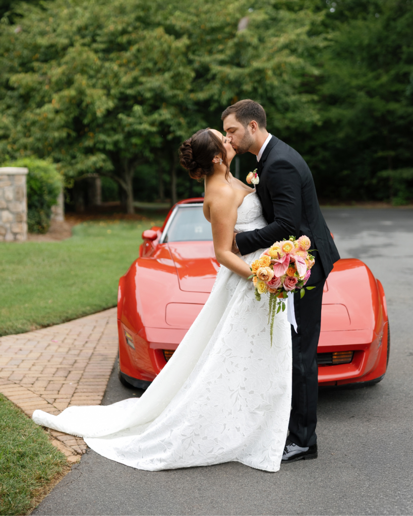 Lauren and Peyton kiss in front of a red vintage car while holding a colorful bridal bouquet after their wedding ceremony.