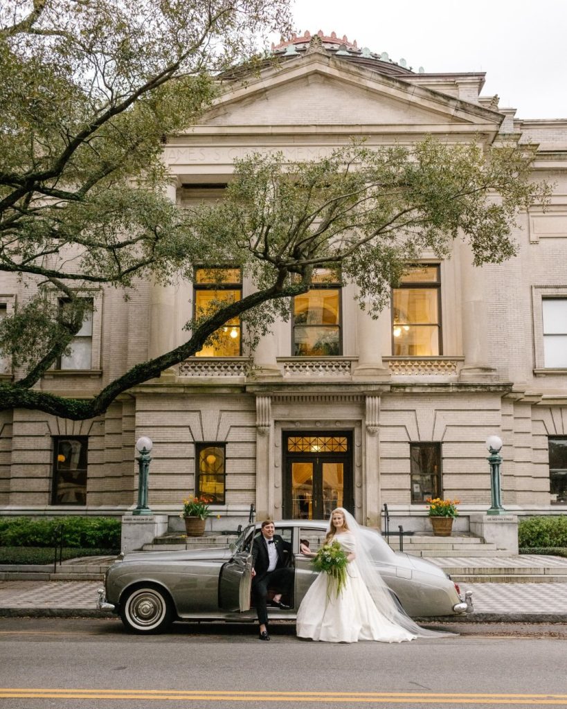 Bride and groom posing beside a vintage convertible outside the Gibbes Museum, a stately stone building with tall windows and an oak tree draped over the entrance.