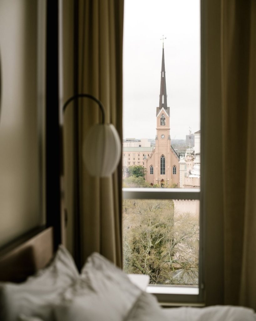 View of a church steeple through a hotel room window, framed by tan curtains with white pillows resting on the bed in the foreground.