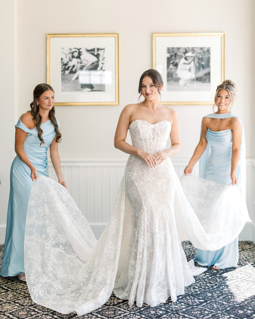 Hannah stands with two bridesmaids in soft blue dresses as they arrange the train of her lace wedding gown.