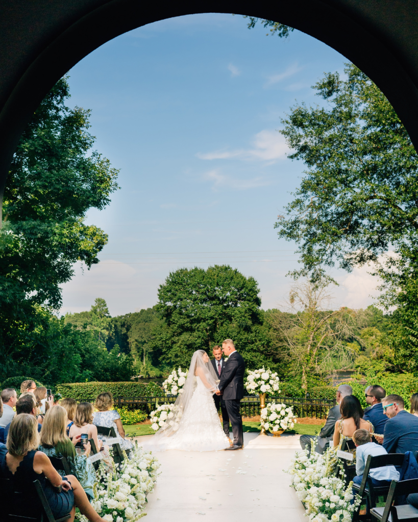 Kelly and Jason stand together beneath an outdoor arch during their garden ceremony at Riverwalk Event Venue, surrounded by white florals and guests.