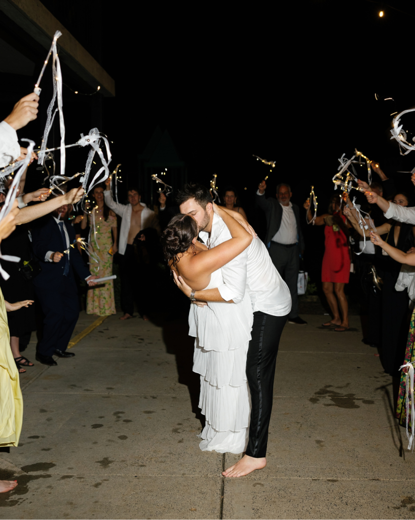 Newlyweds Lauren and Peyton share a kiss during their nighttime wedding exit surrounded by guests waving illuminated ribbon wands.