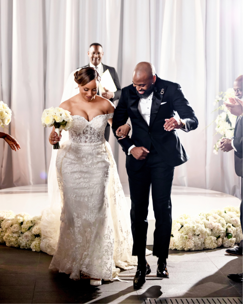 Joy and Edward smile together after their ceremony, with Joy carrying a bouquet of white roses and wearing an embellished off the shoulder wedding gown.