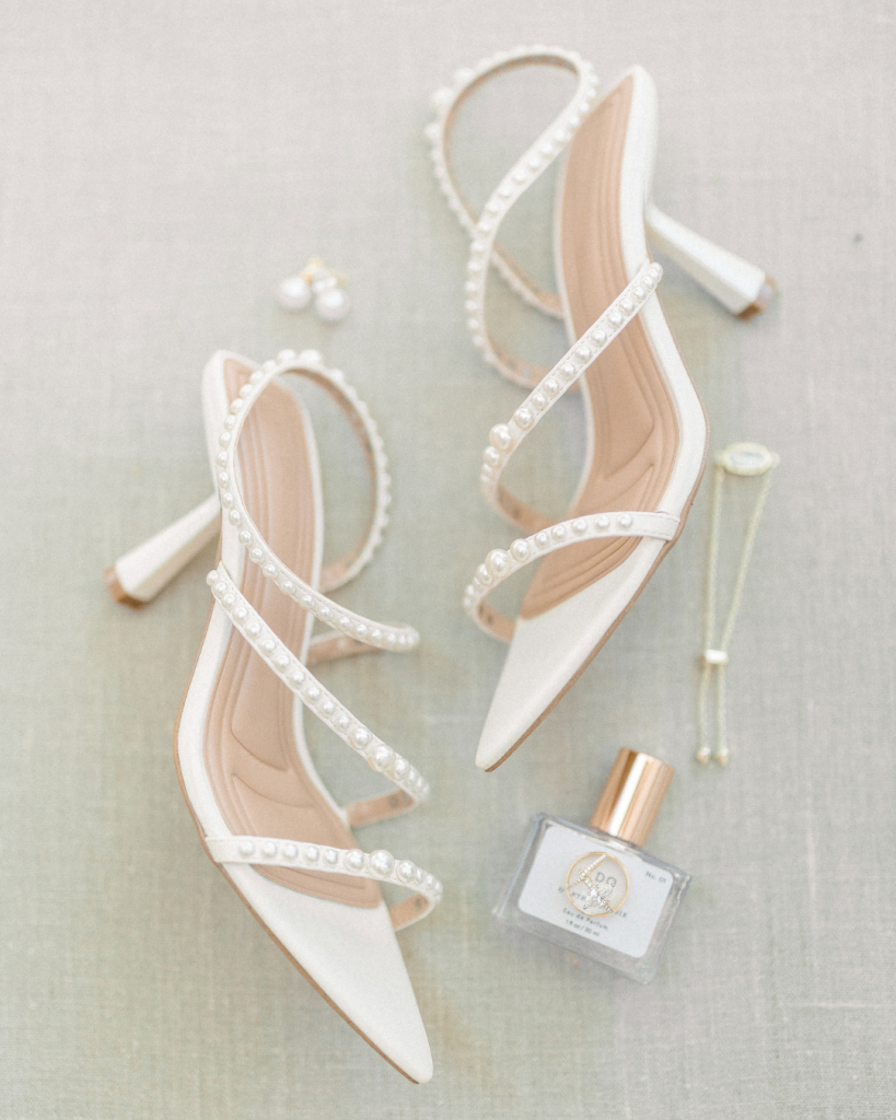 White pointed toe bridal heels embellished with pearls photographed alongside perfume and wedding jewelry.