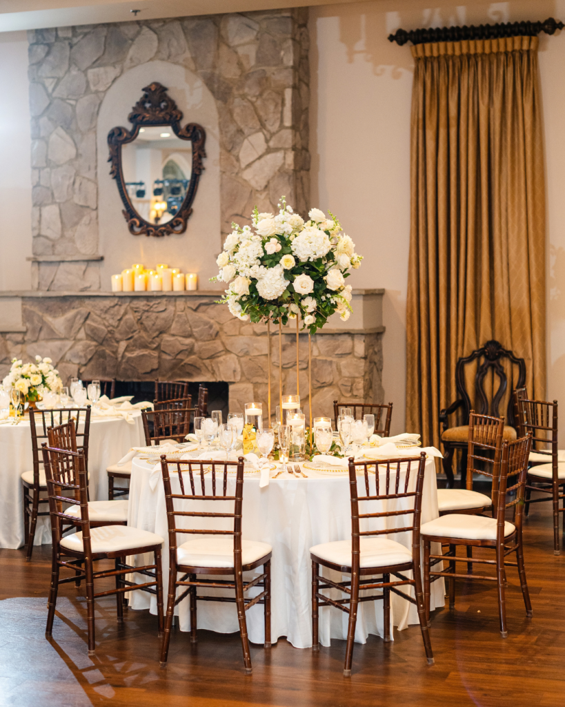Reception table at Riverwalk Event Venue featuring a tall white floral centerpiece, candles, white linens and dark wood chairs beside a stone fireplace.
