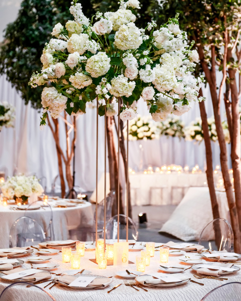 Close view of an elevated white floral centerpiece featuring hydrangeas, roses and greenery above a candlelit reception table.