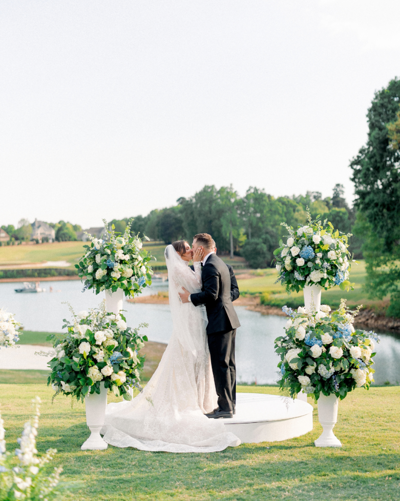 Hannah and Johnathan kiss at their outdoor waterfront wedding ceremony surrounded by large blue and white floral arrangements.