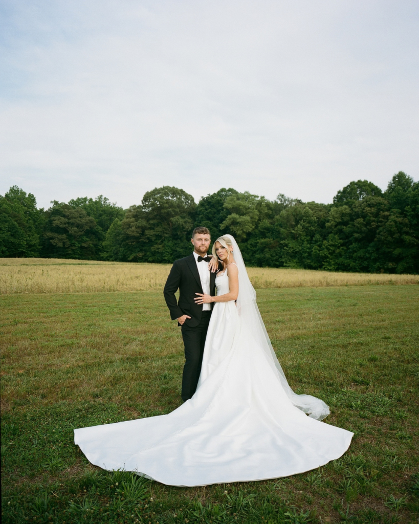Carly and JT walk hand in hand outdoors at The Lumen House, surrounded by green trees and landscaped grounds.