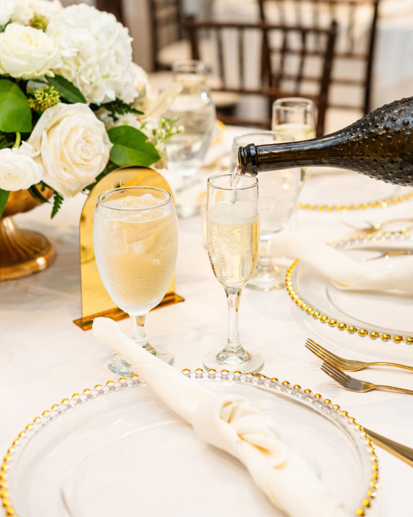 Champagne is poured at a reception table decorated with white roses, gold details, beaded chargers and candlelight at Riverwalk Event Venue.