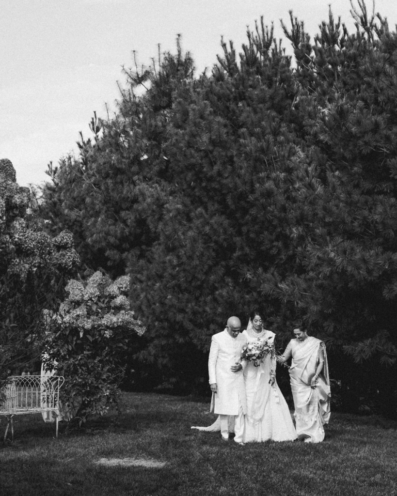 black and white photo of mom and dad walking bride down the aisle near treeline outdoor wedding ceremony south asian jewish multicultural