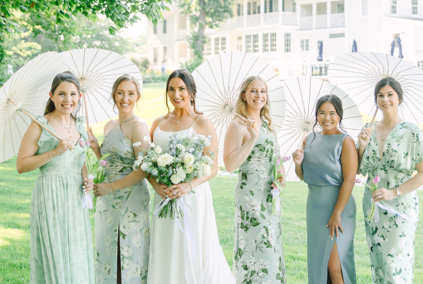 Bride and bridesmaids carrying white parasols in mismatched pale blue, sage and floral-print dresses outside Kent Island Resort.