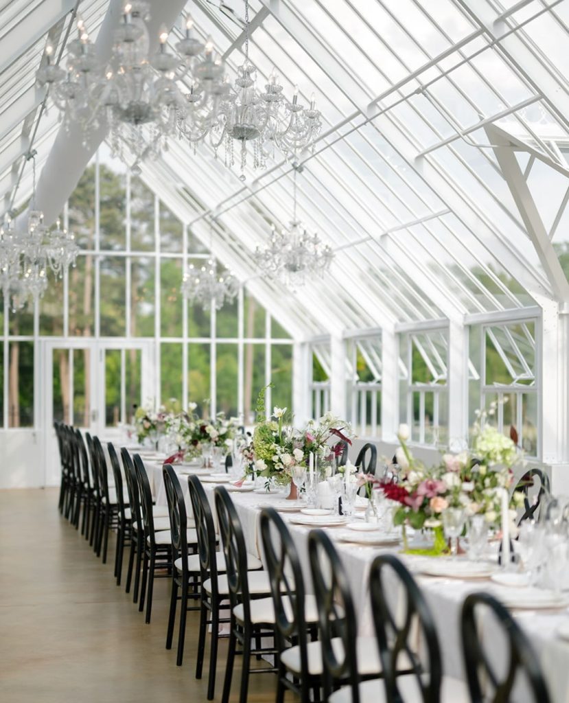 Long reception table with black chairs and floral centerpieces inside a glass conservatory with chandeliers