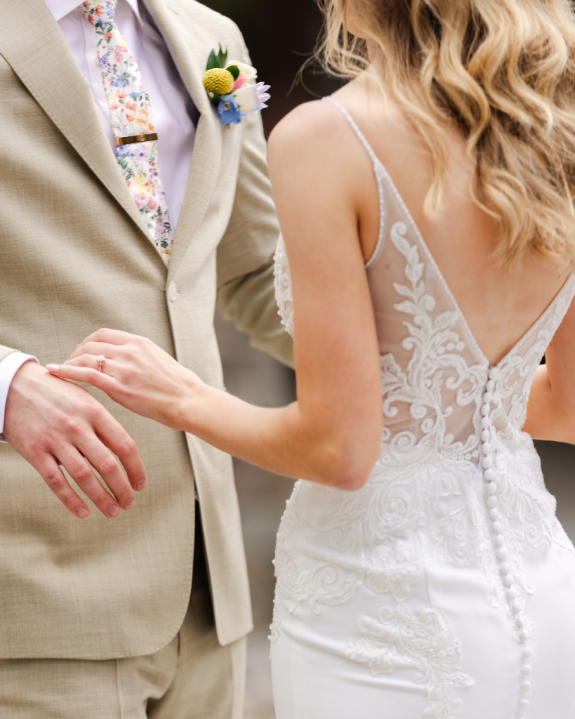 Close-up of a bride in a lace wedding gown touching the groom’s wrist as he wears a tan suit, floral tie and colorful boutonniere.