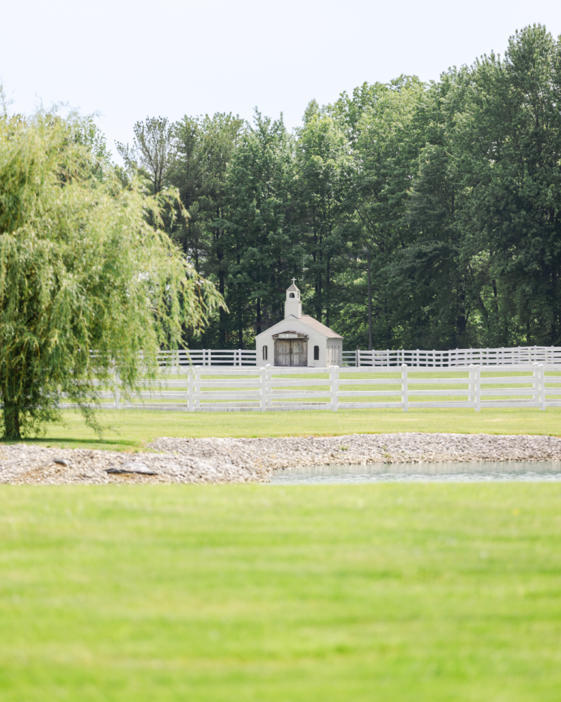 Little Chapel on the Prairie at The Friesian Empire viewed across a green lawn, white fencing and pond.
