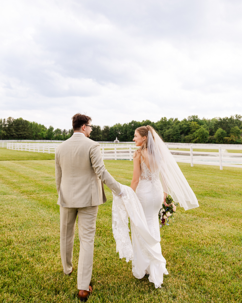 Bride and groom walk hand-in-hand across the lawn at The Friesian Empire beside white fencing and a pond.