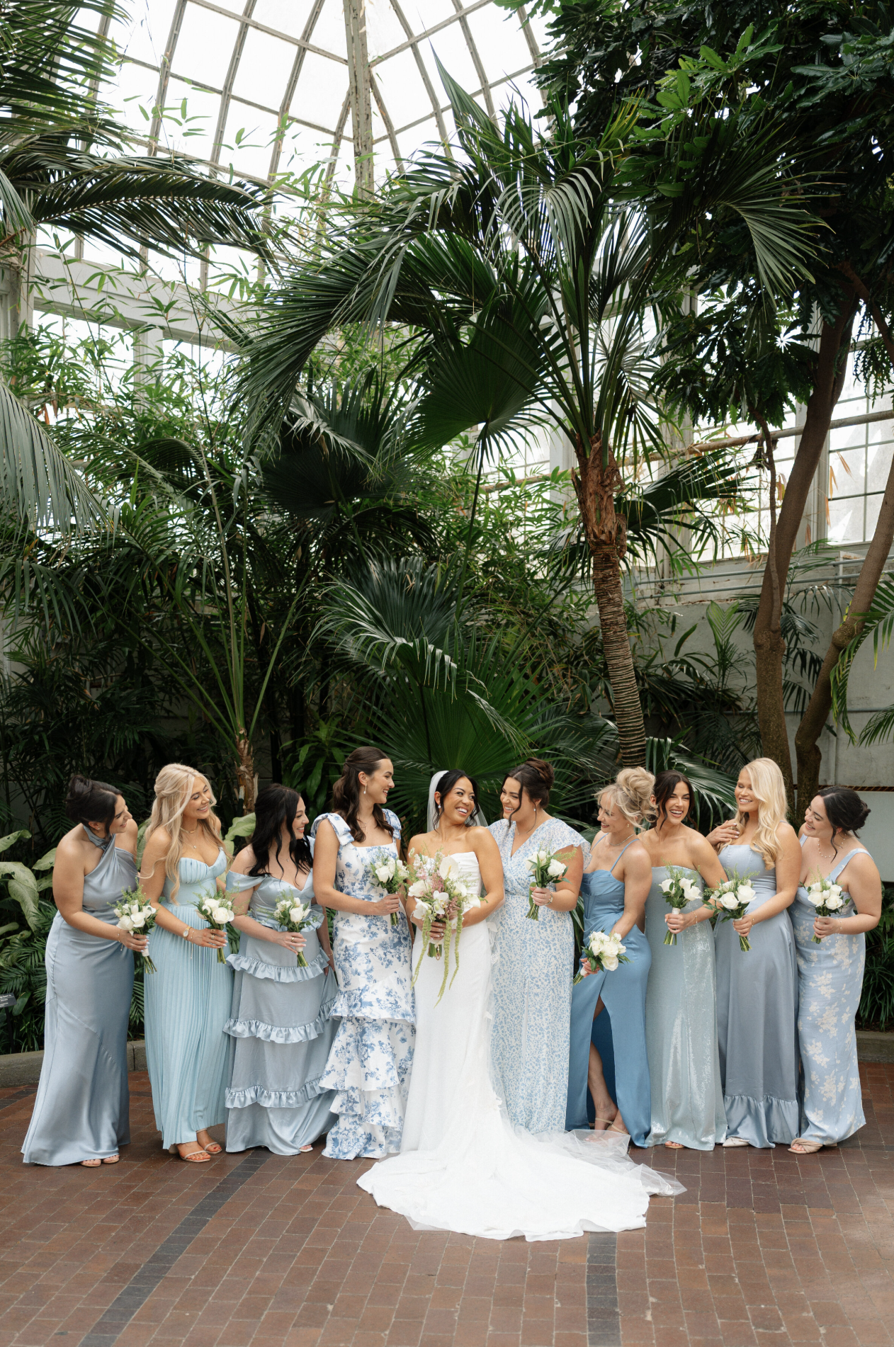 Bride and wedding party posing beneath palm trees inside a conservatory.