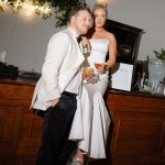 Couple posing together at their wedding reception, holding drinks beside a wooden bar.