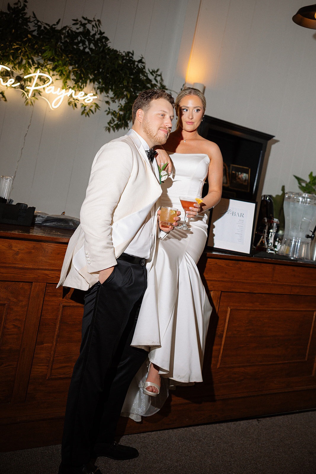 Couple posing together at their wedding reception, holding drinks beside a wooden bar.