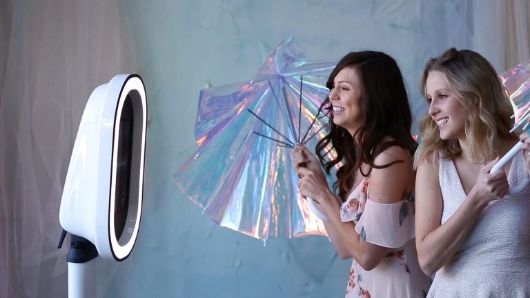Two wedding guests pose with iridescent umbrellas in front of a Best Booths in Town ring-light photo booth.